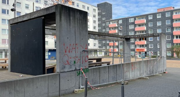 Die Betonkonstruktion des Gemeinschaftspavillons Brunnenplatz 1. Die Flächen sind teils mit roten Graffiti Schriftzügen bemalt. Am Rand steht zur Absicherung am Gefälle ein Bauzaun, der zusätzlich mit rotem Absperrband gekennzeichnet ist.